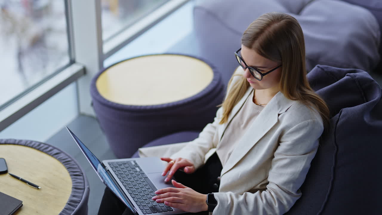 Calm relaxed young business lady working on laptop sitting in a chair. Freelance working lady in a comfortable place near big windows.