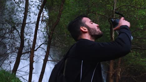 Young-thirsty-male-drinking-water-in-front-of-a-waterfall,-slow-motion-medium-shot