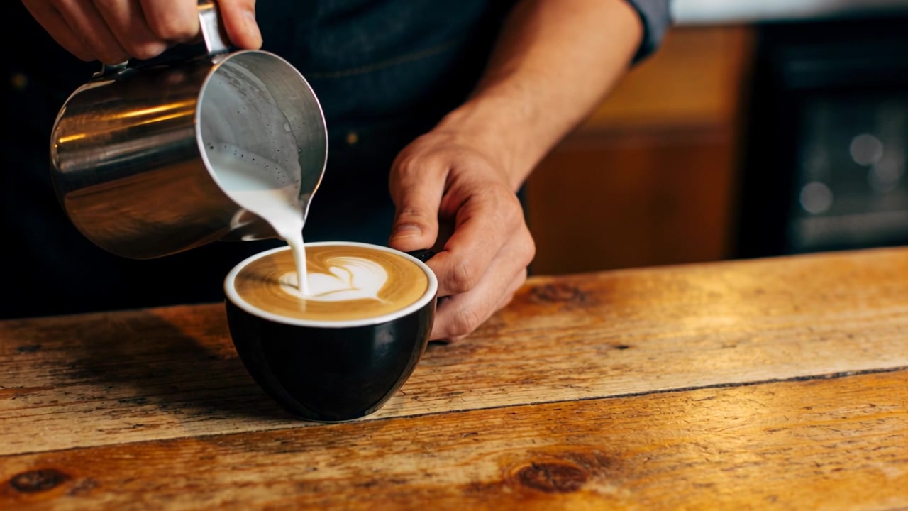 Barista con maglia scura che versa il latte, decora una tazza con un cuore e poi una foglia al bancone con una lattiera.