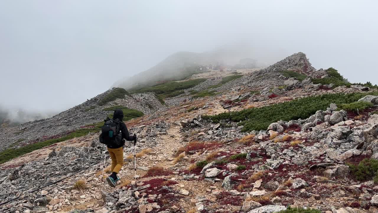 Un escursionista solitario attraversa un crinale roccioso nelle Alpi Giapponesi Settentrionali. La nebbia avvolge le imponenti montagne, creando un'atmosfera autunnale suggestiva. Catturato in formato paesaggio