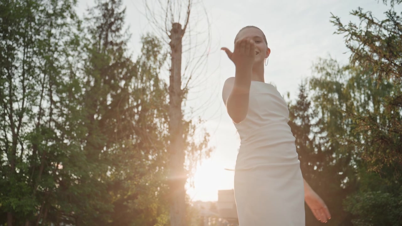 Young woman in white dress performs graceful dance movements outdoors on wooden planks with sunlight filtering through trees. Her arms rise and fall her hair moves naturally as woman poses