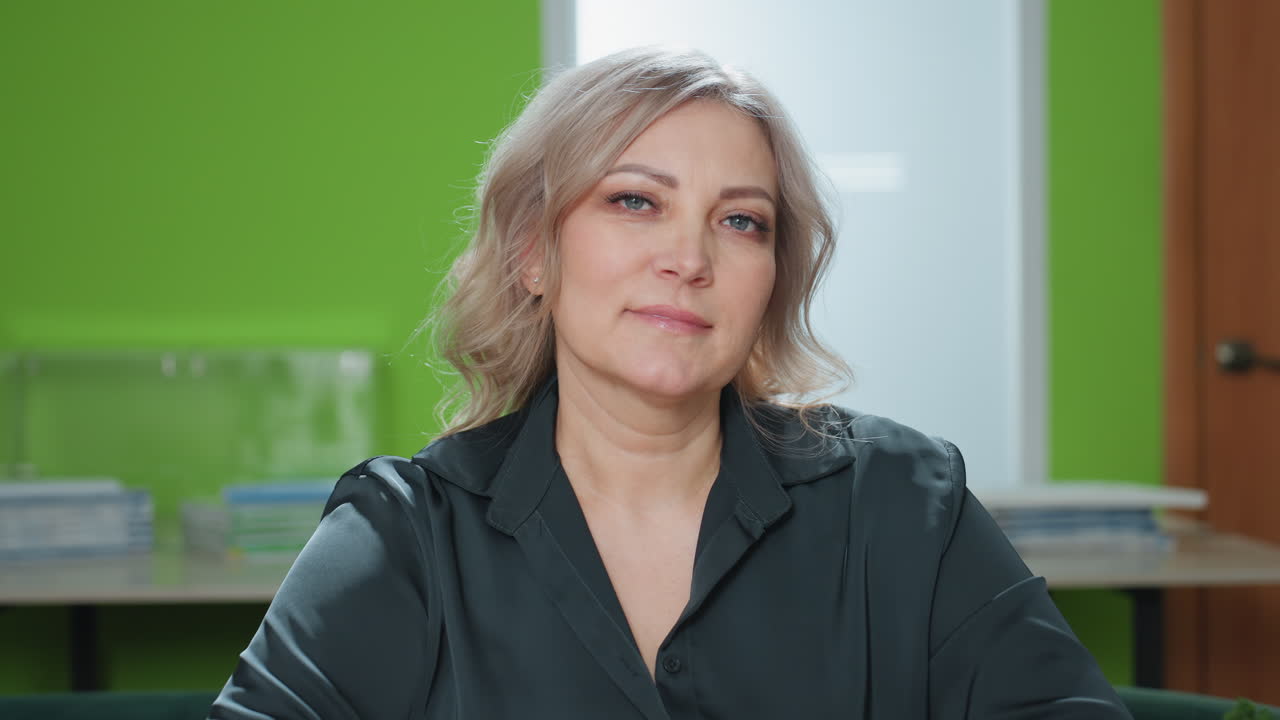 woman in professional attire seated at desk with hands resting on closed notebook, tilting head slightly with confident expression in modern office with green accent wall and architectural model
