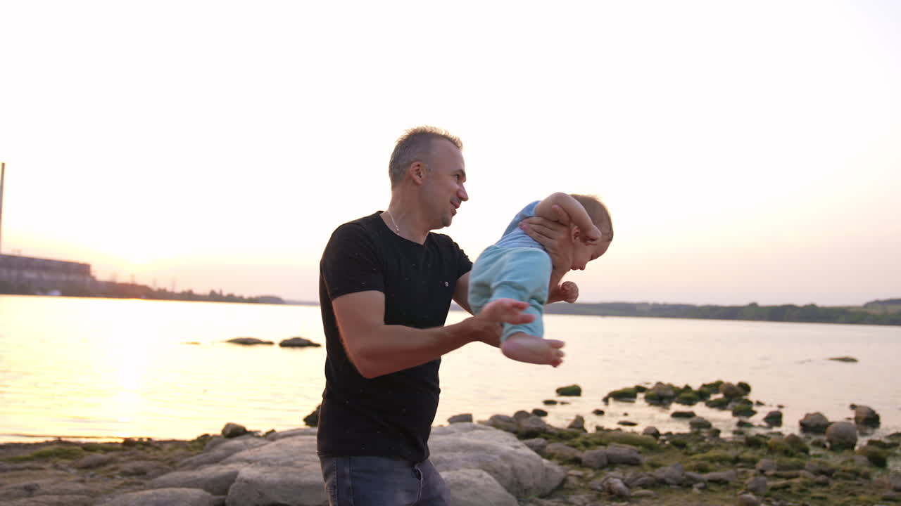 Man in black t-shirt whirling little baby. Father playing with cute adorable son who's laughing cheerfully. River at backdrop.