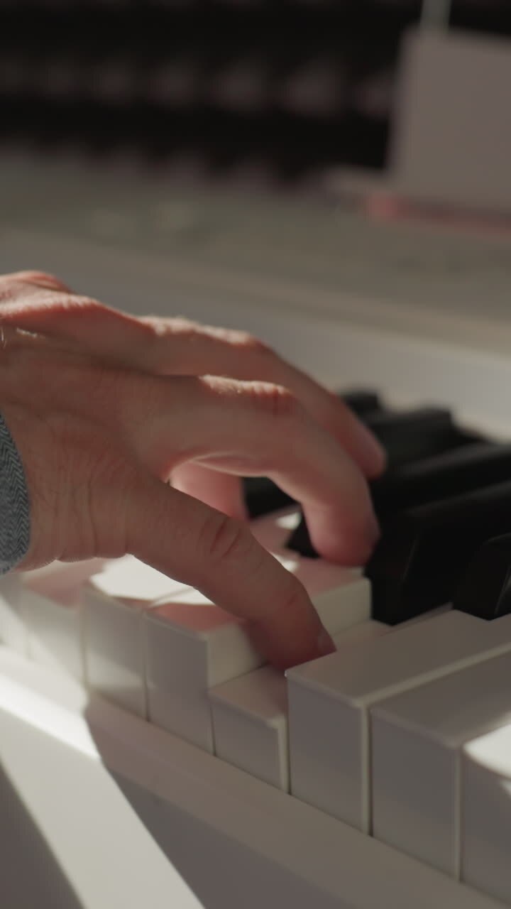 Close-up view of a hand in a blue sleeve shirt gracefully playing the piano keys, with a softly blurred background. The focus on the hand captures the delicate motion and expression of music