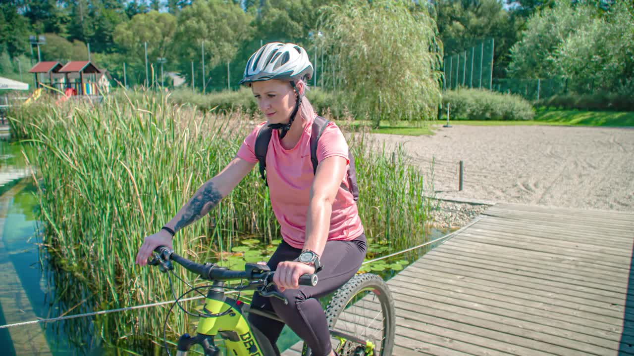 Adult Couple Cycling Across Wooden Bridge Past Water Lilies At Radlje Ob Dravi Park In Slovenia. Slow Motion