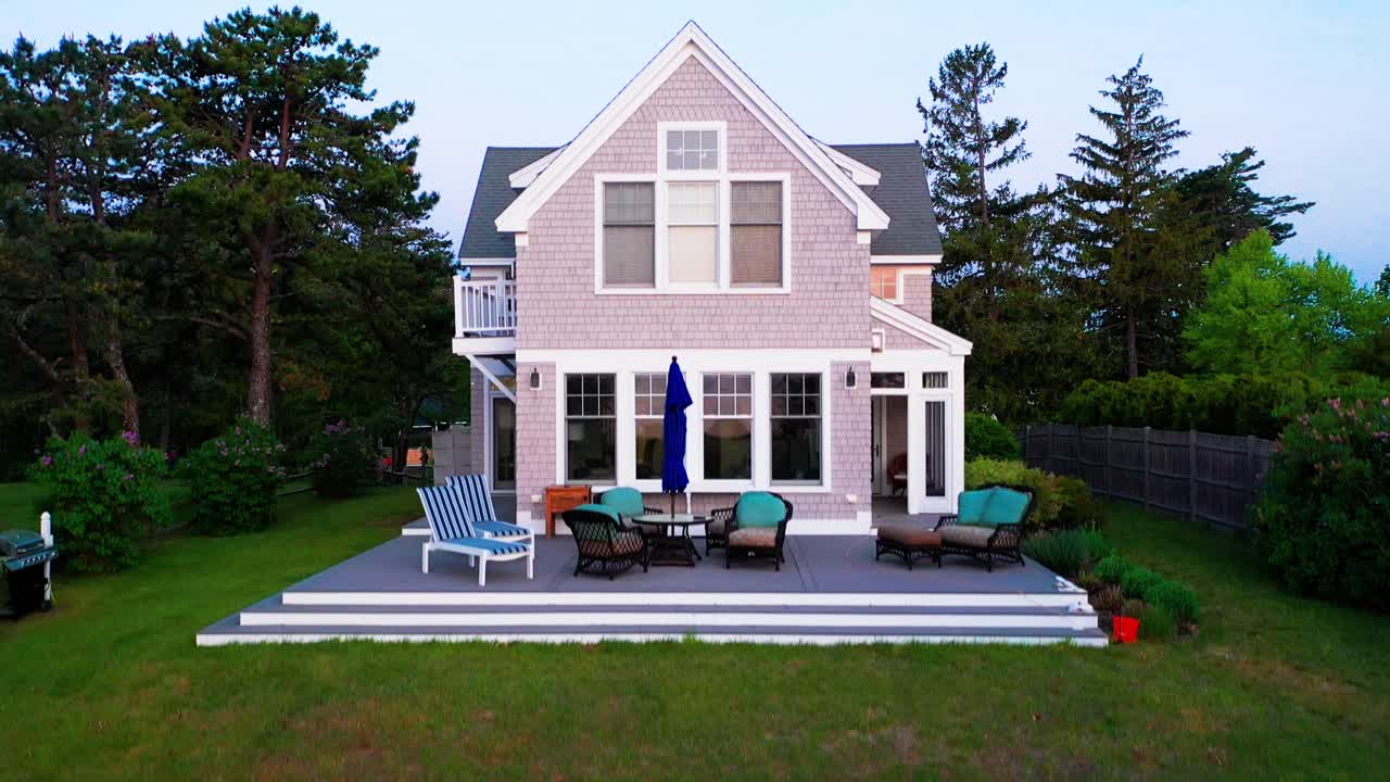 Aerial drone real estate video showcasing a charming suburban cottage-style house with a spacious back porch, cozy patio furniture, manicured lawn, and surrounded by tall, lush green trees