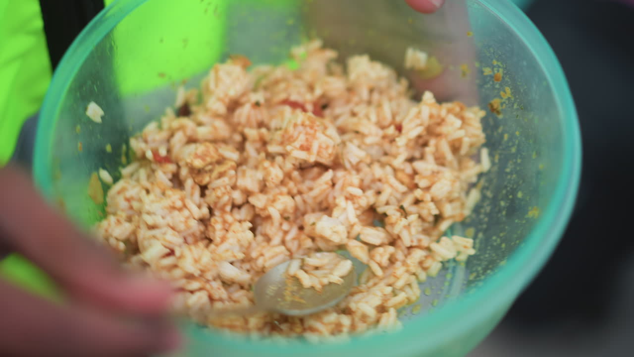 Close-up of hands holding green plastic bowl filled with seasoned rice, person eating with spoon, visible warm tones from spices, casual outdoor meal suggesting homemade comforting food during break time