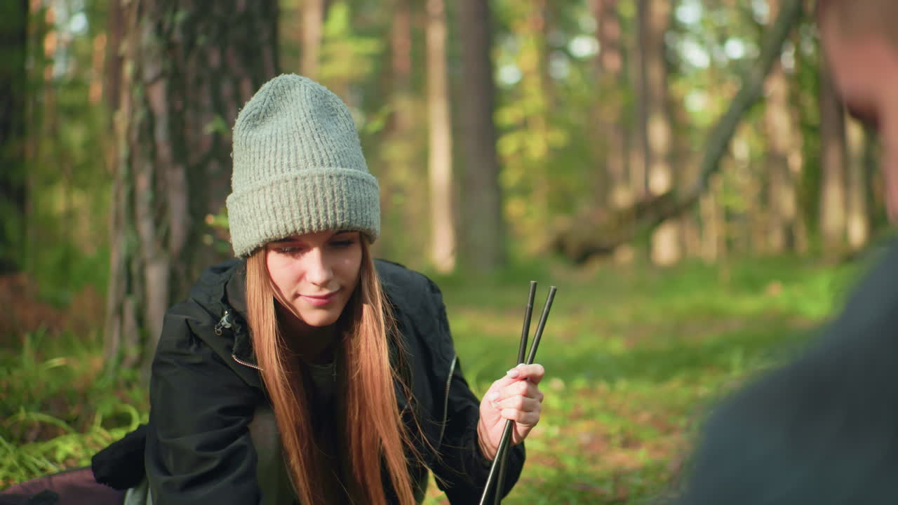 Lady holding camping tent poles in hand while playfully handing one to companion who prepares tent setup in forest, smiling and enjoying peaceful moment surrounded by nature and sunlit trees
