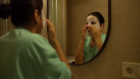 una chica morena confiada con piel marrón en una camiseta verde hace una máscara especial aplicada a su cara por la noche en un apartamento moderno en el baño antes de ir a la cama