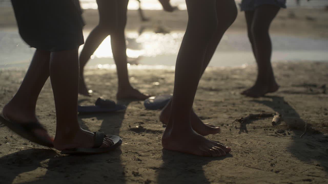 group of kids excitedly pick up their flip flops and run through the shallow water at the edge of the sea, laughing and splashing as they go. sun reflexions on background.