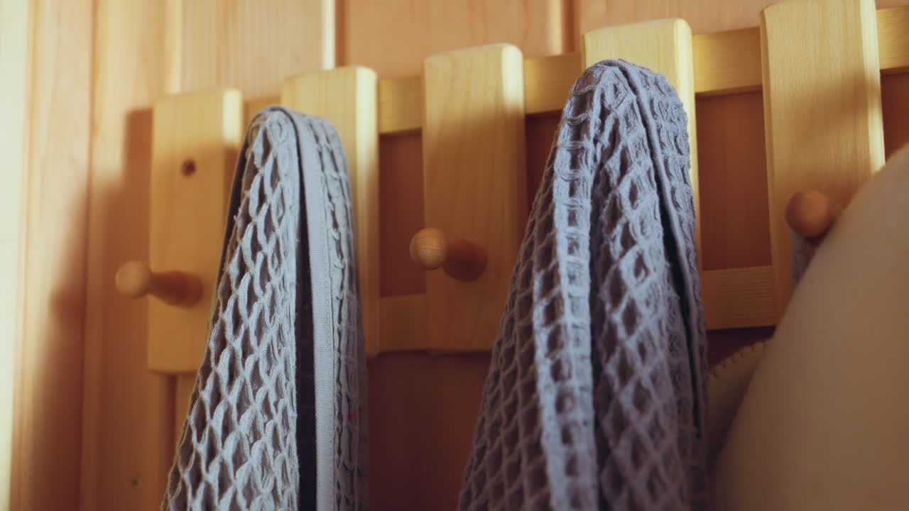 close up of sauna corner with wooden hook rail supporting hanging waffle weave towels and felt hats, warm sunlight casting gentle shadows on pine panels