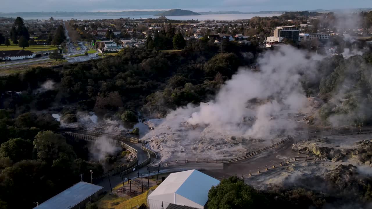 Beautiful aerial over Te Puia&rsquo;s Pohutu Geyser reveal of scenery Rotorua Lake on horizon during golden hour