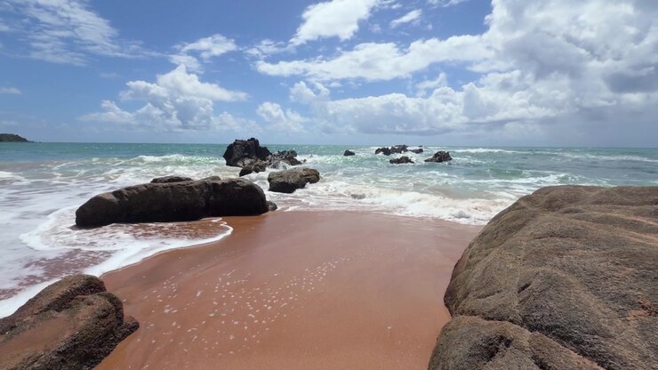 Vista panorâmica de ondas do oceano quebrando em uma praia tropical de areia com grandes formações rochosas escuras sob um céu parcialmente nublado em Porto Rico.