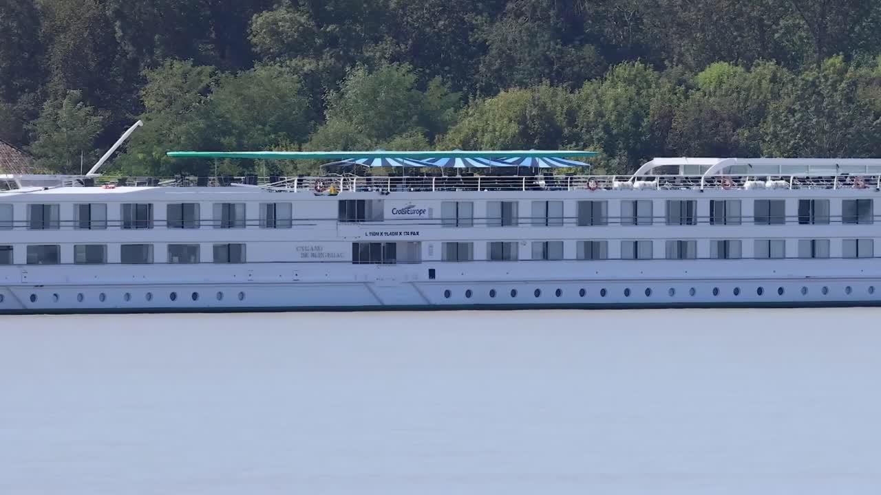 A white cruise ship sails smoothly along a river, bordered by dense greenery.