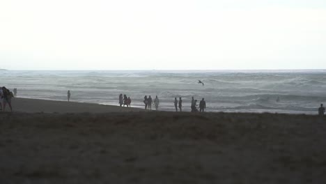 Tiro-Estático-De-Bajo-Nivel-Que-Captura-La-Silueta-De-La-Gente-En-La-Playa-De-Arena-Con-Olas-Rápidas-Rompiendo-La-Orilla