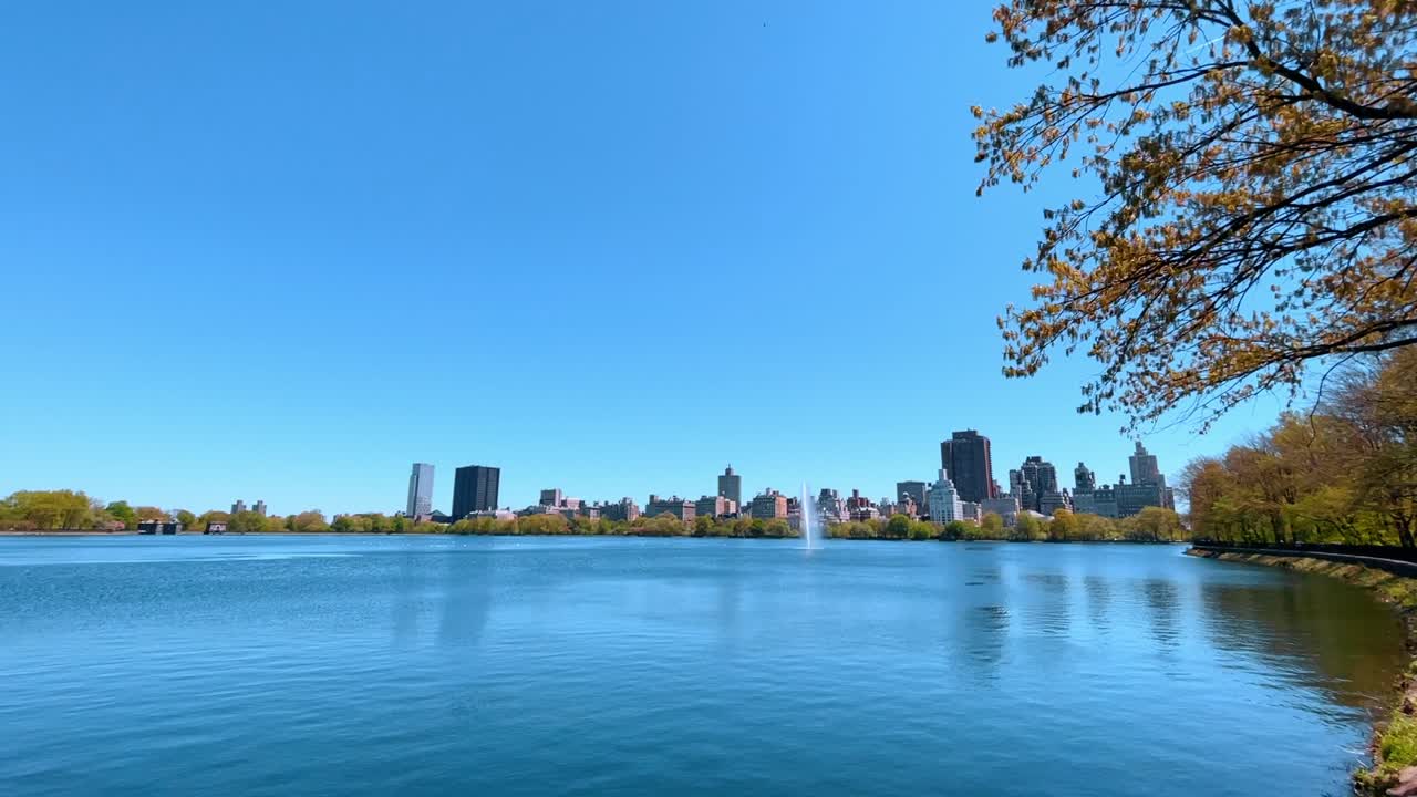 Reservatório do Central Park com fonte e o horizonte de Manhattan ao fundo. Vista ampla da superfície da água, árvores e edifícios da cidade sob céu azul.