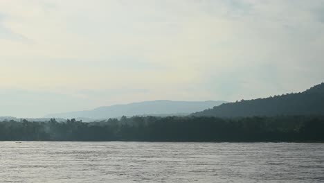 Río-Mekong-Fluyendo,-Se-Ve-Un-Bote-Navegando-Por-El-Agua-Que-Fluye-Desde-La-Izquierda-Del-Marco-Yendo-Hacia-La-Derecha-Mientras-Se-Ven-Nubes-Nebulosas-En-El-Horizonte-Y-Montañas-Boscosas