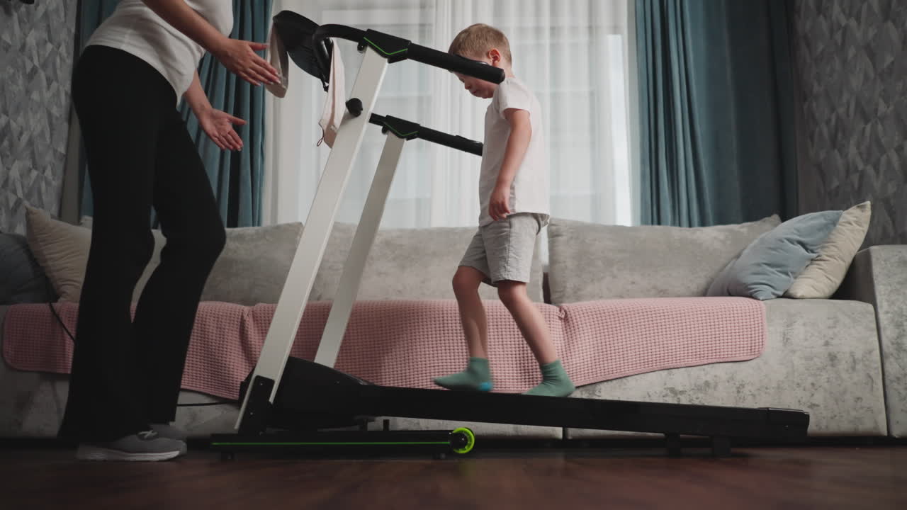 Female trainer in white top and black pants assists young boy in white shirt and green socks on treadmill, providing guidance and encouragement during focused training session