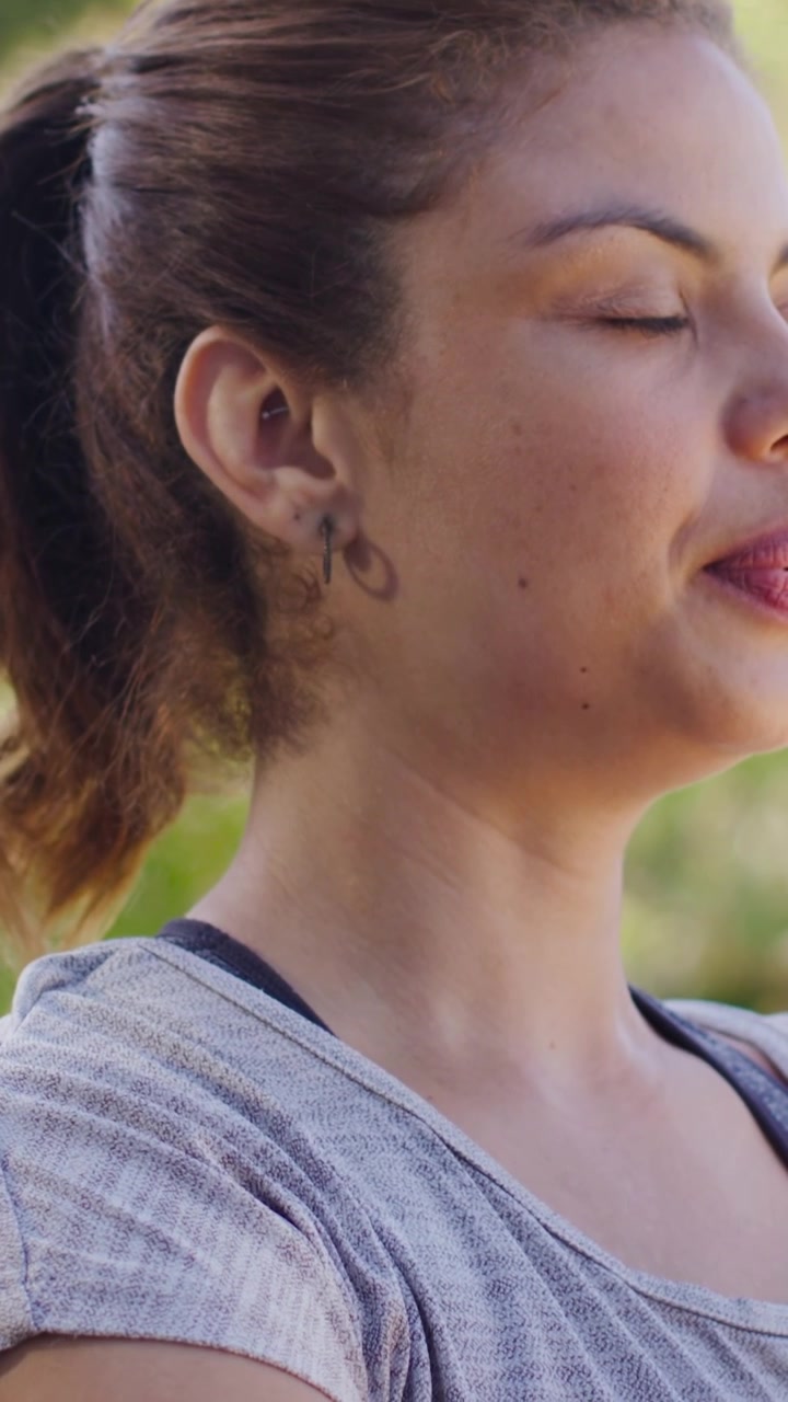 Video verticale: Donna che abbassa il braccio, chiude gli occhi e respira con calma al parco, con maglietta grigia, spazio per il testo.
