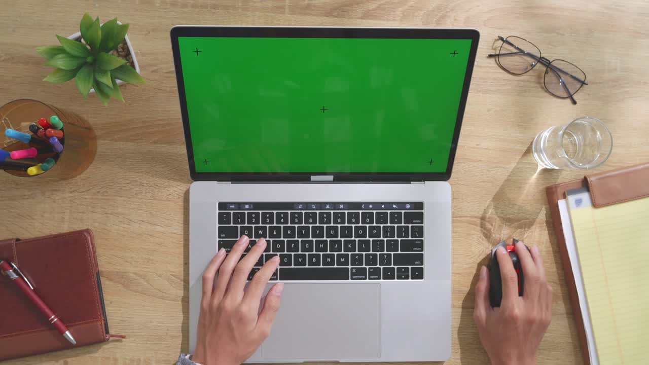 Top down view of a laptop computer with mock up green screen chromakey display on a wooden office desk next to notebook with pens, glasses, and a glass of water. Slow zoom out, close up