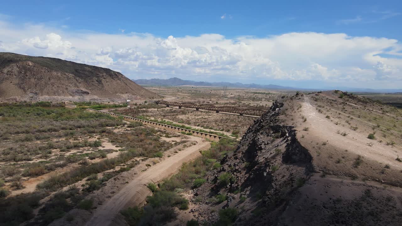 drone panoramización alrededor de un acantilado que revela un viejo puente de acero rústico en un día soleado, cielo azul con grandes nubes blancas presa y puente gillespie, río gila