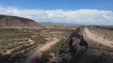 Drone-Panning-around-a-cliff-revealing-an-old-rustic-steel-bridge-on-a-sunny-day-,-blue-sky-with-large-white-clouds-Gillespie-Dam-and-Bridge,-Gila-river