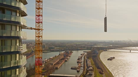 Roter-Und-Gelber-Baukran,-Der-An-Einem-Wolkenkratzer-Auf-Der-Baustelle-Befestigt-Ist,-Hebt-Die-Last-Und-überträgt-Sie-An-Einem-Sonnigen-Tag-Auf-Das-Dach-Des-Gebäudes