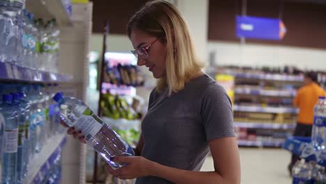 young mother is choosing a bottle of water in beverages department in the supermarket, while her little baby is sitting in a