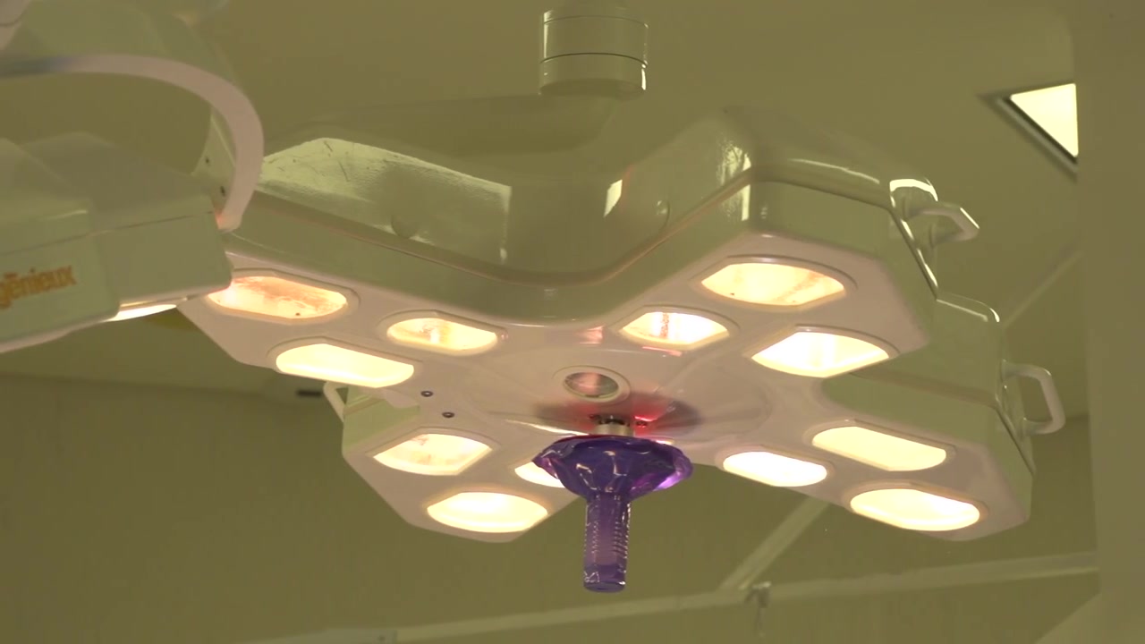 Primo piano con inquadratura dal basso di una lampada chirurgica a più bulbi accesa in una sala operatoria, che mostra una copertura sterile viola per la maniglia ed emette una luce calda e brillante durante una procedura ospedaliera.