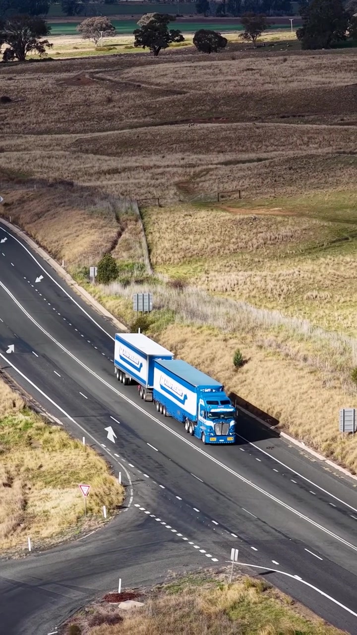 Un autoarticolato blu con doppi rimorchi viaggia lungo una strada rurale attraverso una campagna arida.