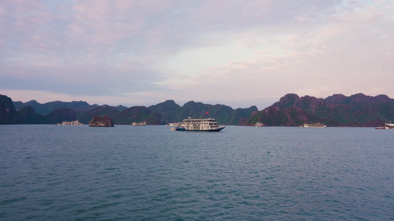 Several tourist cruise ships navigate through the tranquil waters of Ha Long Bay, surrounded by dramatic limestone karst formations rising from the sea against a soft pastel sunset sky