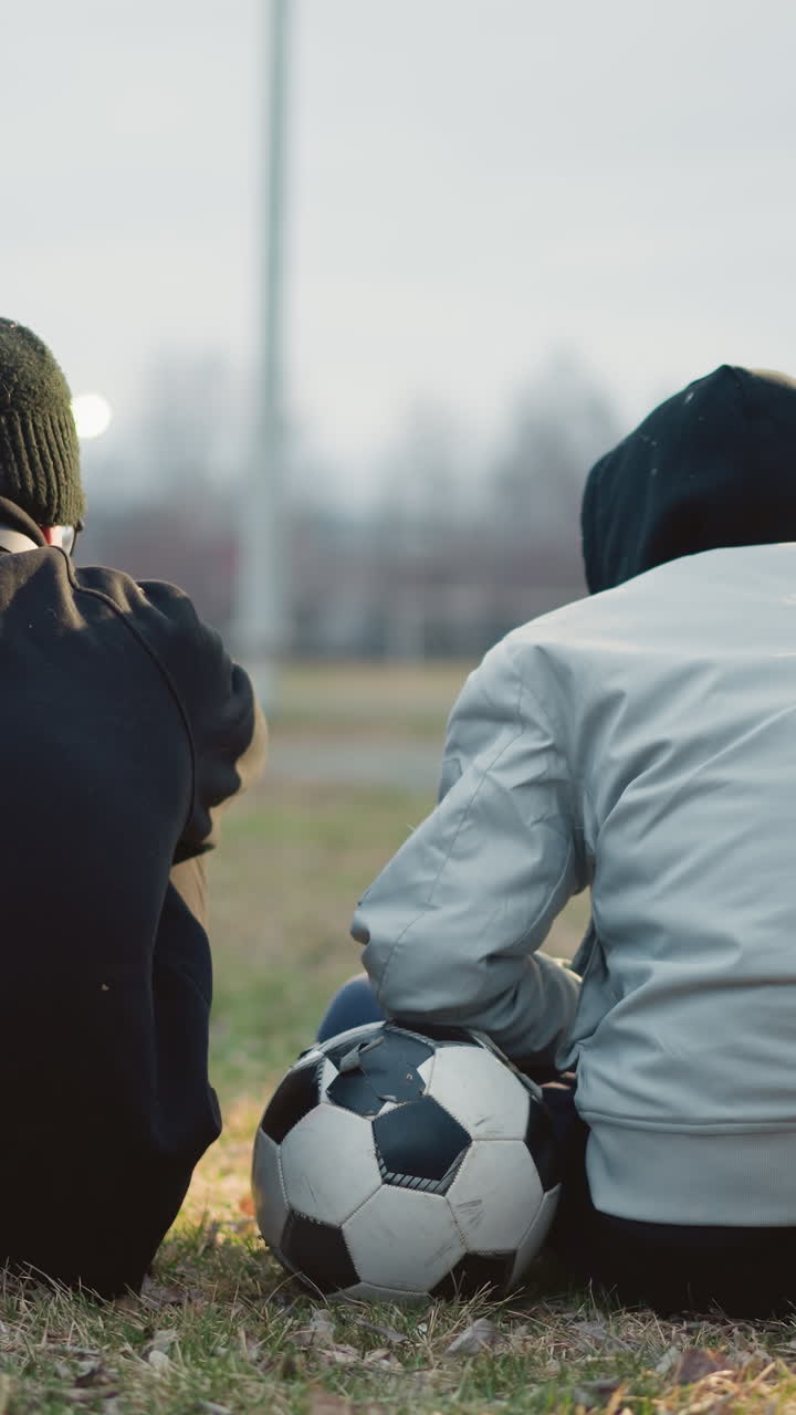 primer plano de dos personas sentadas en un campo de hierba, con uno ajustando su mano en una pelota de fútbol, durante el crepúsculo, con luces de fondo borrosas