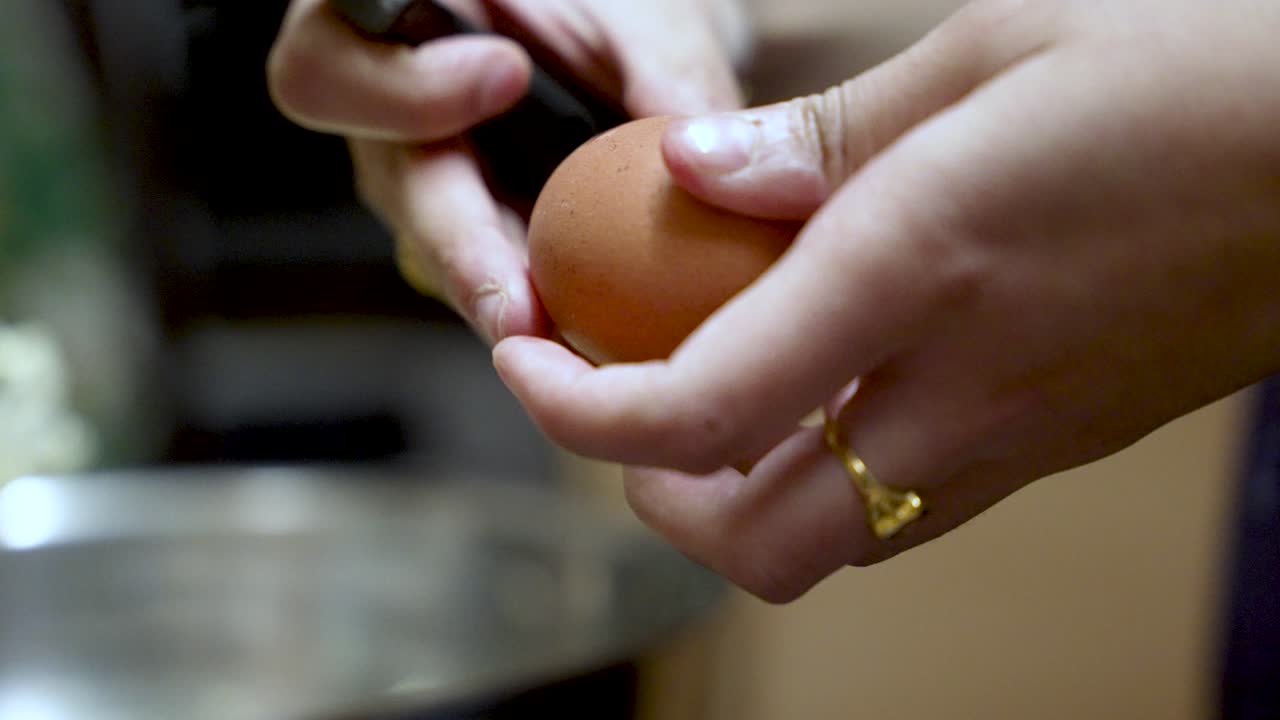 A hand cracks a brown egg using a knife over a bowl in a kitchen. slow motion