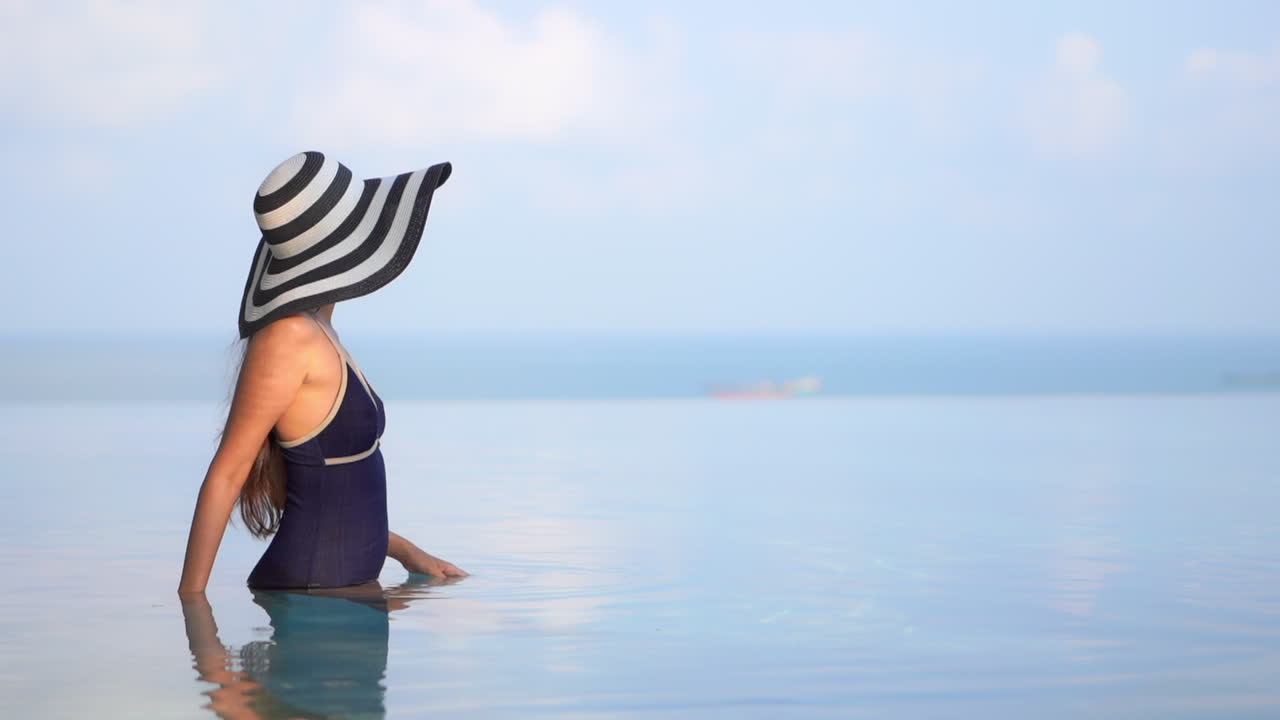 Beautiful woman in dark bathing suit and matching hat stands in calm infinity swimming pool overlooking a tropical island beach. Luxury travel vacation concept. Tanning relaxation and paradise holiday