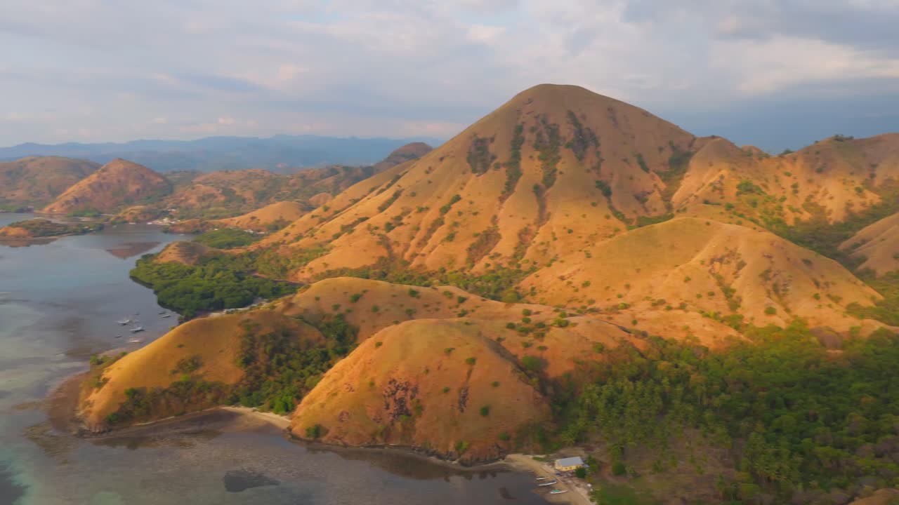 Aerial view of rugged golden hills meeting turquoise shallows with scattered boats in Komodo National Park