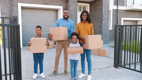 portrait of happy african american family with small children standing at new house at suburb and smiling with carton boxes in hands