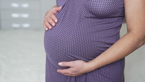 expectant lady standing half-turned in her bedroom by the bed. woman expecting a baby wearing a nightgown and stroking her belly. close up.