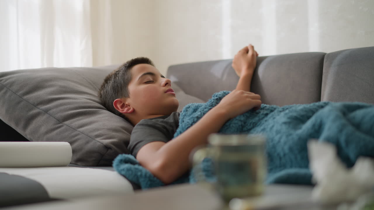 Sick kid lying on couch covered in blanket, coughing while resting hand on his head, tissue nearby and blurred view of cup and tissue on table