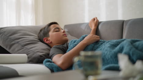 sick kid lying on couch covered in blanket, coughing while resting hand on his head, tissue nearby and blurred view of cup and tissue on table