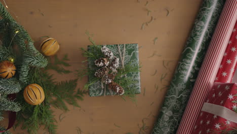 top view of the decorator&#039;s desktop Fully visible the table with the decorations. Female hands put and finalize Christmas gift wrapped in craftool paper on a wooden table