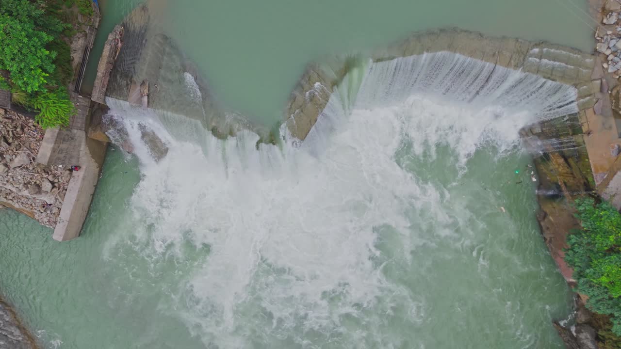 Aerial drone view of a beautiful waterfall merging into a flowing river, capturing the natural beauty of water movement, rocky terrain, and pristine landscape surrounded by greenery