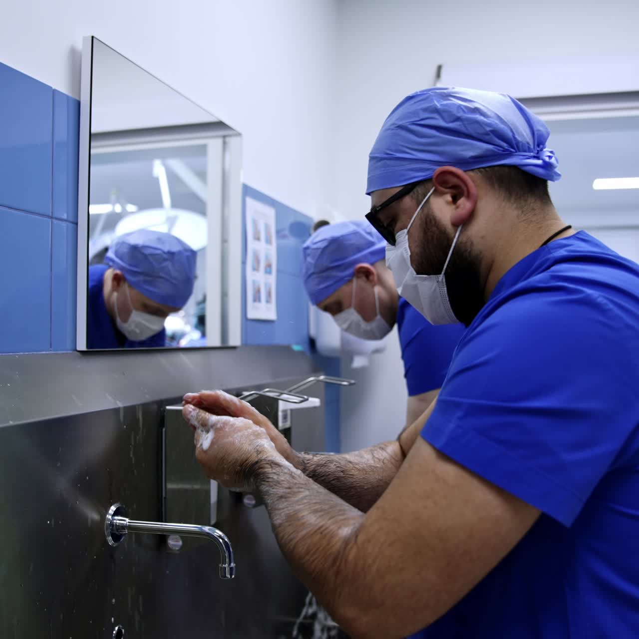 Two male surgeons wearing blue uniforms and masks are in the pre-operational room. Doctors wash hands carefully before the surgery