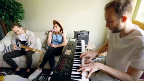 young bearded keyboardist is playing keyboard while his friends are singing and playing guitar sitting on sofa during rehearsal at home. close shot of hands on key buttons.