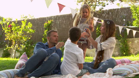 Familie-Sitzt-Auf-Einer-Decke-Im-Garten-Und-Bläst-Seifenblasen