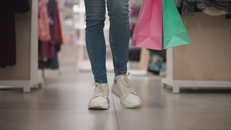 leg view of shopper walking confidently through retail clothing store aisle carrying vibrant pink and green shopping bags, in a modern, neatly arranged retail environment