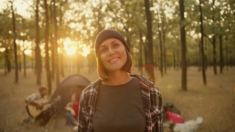 Portrait-of-a-happy-girl-with-a-bob-hairstyle-in-a-black-hat-in-a-plaid-shirt-against-the-backdrop-of-hikers-and-tents-in-a-Sunny-summer-green-forest.-Happy-woman-posing-and-looking-at-camera-while-hiking