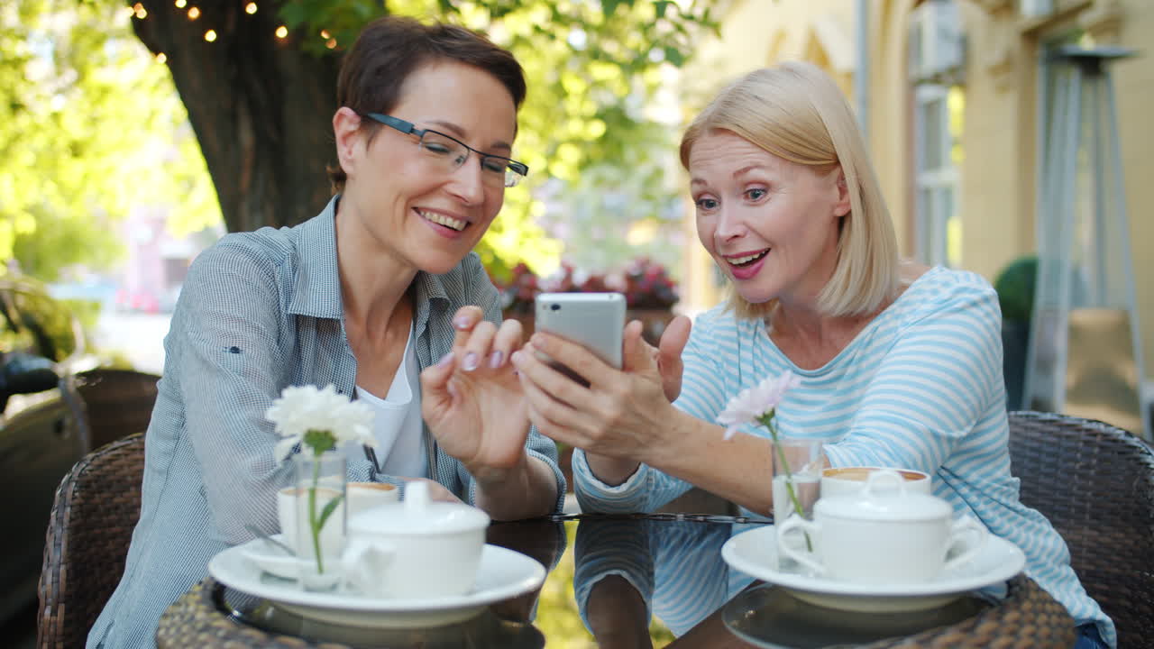 Two women friends enjoying coffee and looking at a smartphone in a cafe
