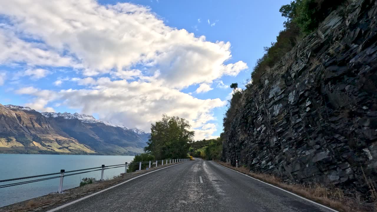 Car travels along winding lakeside road, cliffs and mountains visible, daylight, smooth forward camera movement