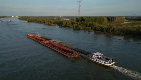 Luftbild--Und-Schwenkaufnahmen-Eines-Langen-Frachtschiffs,-Das-Schüttgut-In-Lastkähnen-Auf-Dem-Fluss-In-Flacher-Landschaft-Transportiert