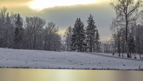 Timelapse-shot-of-white-snow-covered-lakeside-on-a-cloudy-cold-winter-evening-beside-a-road-with-car-passing-by